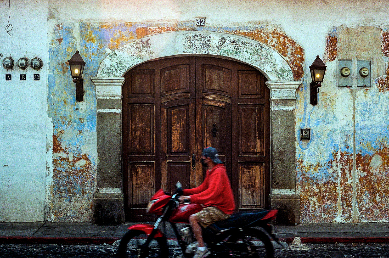 Person on red motorcycle passing by weathered blue wall with wooden door